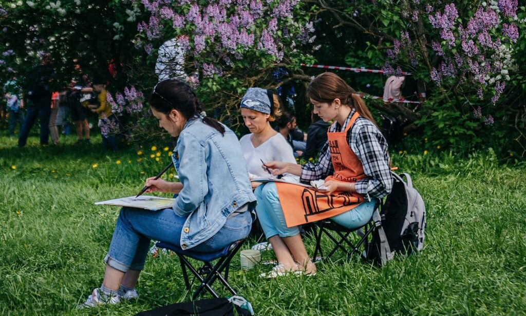 A group of three women are sitting on a lawn creating artwork with lilac trees in the background.