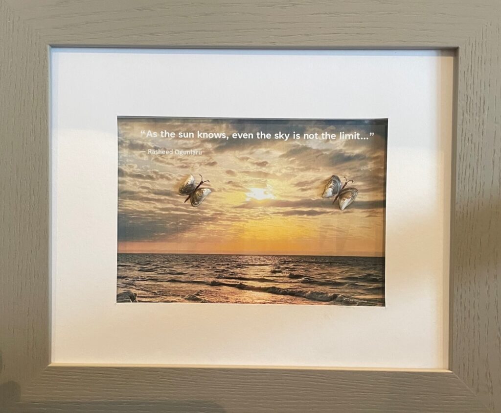 Framed image of a sunset reflecting on the water, with two butterflies made from shells and this quote: As the sun knows, even the sky is note the limit.