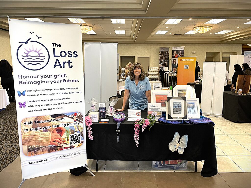 Julie stands with her trade show booth, featuring gifts for sale, draw prizes, decorations and a large banner with this text: The Loss Art. Honour your grief. Reimagine your future. Visit TheLossArt.com to begin your healing journey.